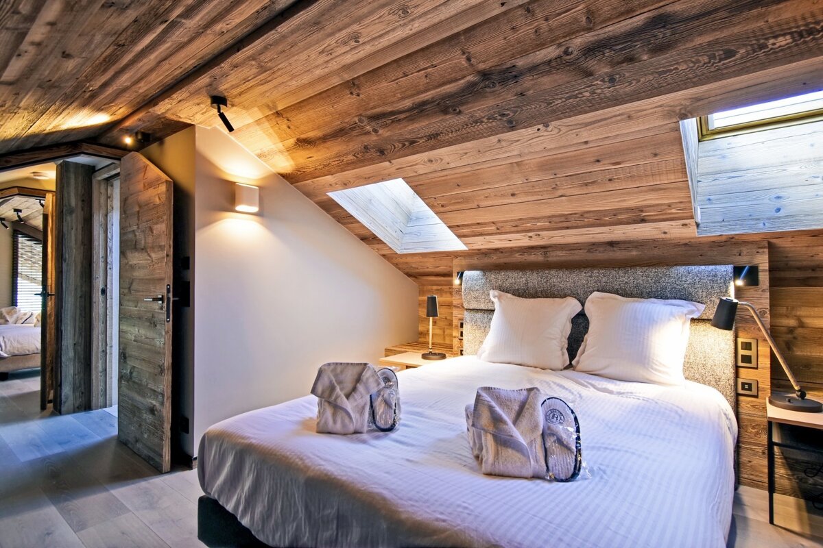 A cozy bedroom features a slanted wooden ceiling, a skylight, and a white bed with robes and slippers. Modern lights illuminate rustic wood walls.
