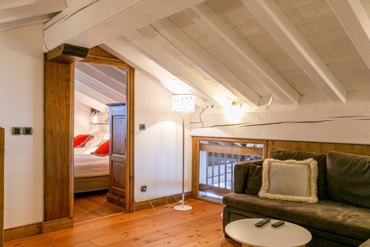 Cozy attic living room with a sloped beamed ceiling, wooden floors, brown sofa, and an open doorway leading to a bedroom with red pillows.