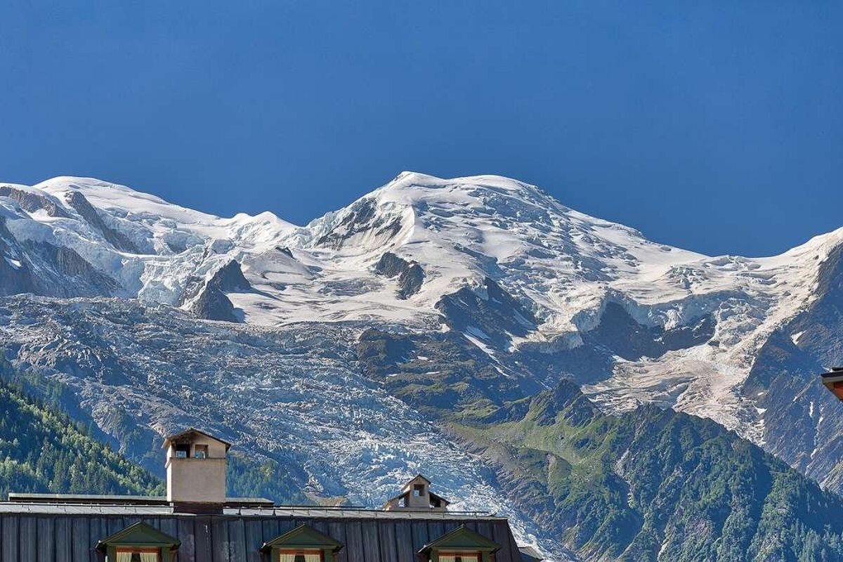 Majestic snow-capped mountain peaks and a glacier tower over alpine rooftops under a clear blue sky.