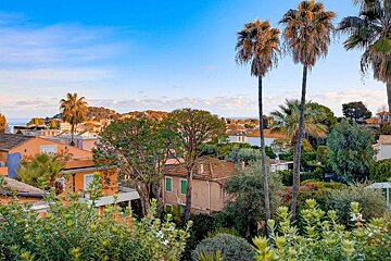 View of a vibrant coastal town with colorful houses, lush green foliage, tall palm trees, and the ocean visible under a clear blue sky.