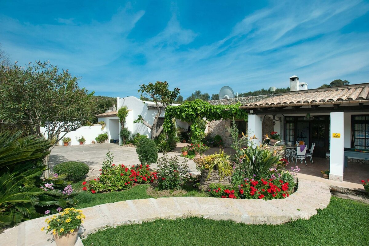 A beautiful white house with a terracotta roof, surrounded by a lush green garden with vibrant red flowers and a stone path under a bright blue sky.