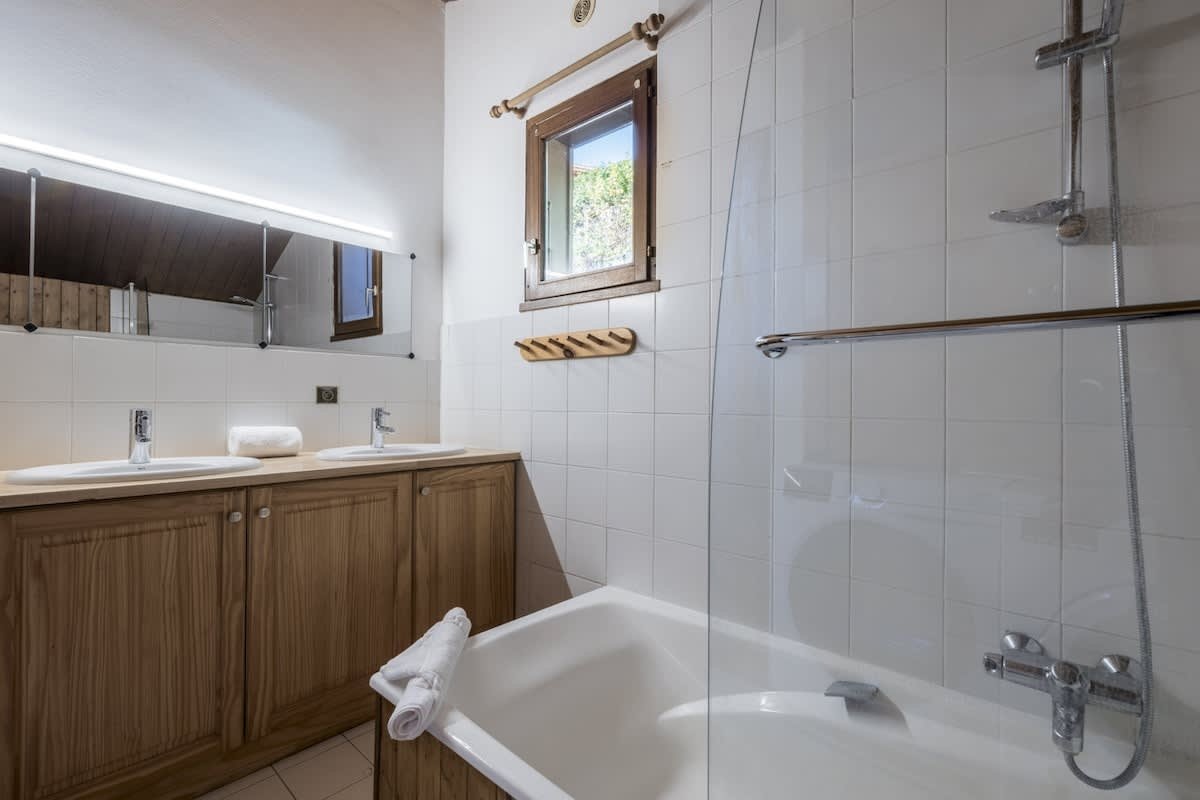 A clean bathroom featuring a wooden double vanity, white tiled walls, and a bathtub with a glass shower screen.