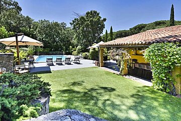 A luxurious villa with a tiled roof overlooks a sparkling pool and a lush green lawn, surrounded by trees under a bright blue sky.