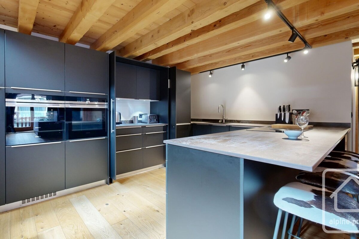 A sleek, modern kitchen with dark grey cabinets, light wooden floors, exposed ceiling beams, and an island with distinctive cowhide stools.