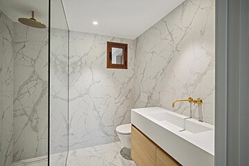 A modern bathroom features white marble walls/floor, gold fixtures, a sleek double sink vanity with a wood cabinet, and a shower area.