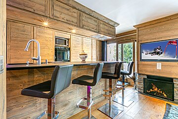 Wooden chalet kitchen/bar with black stools, built-in oven, TV showing skiing, and a fireplace below. Rustic modern comfort.