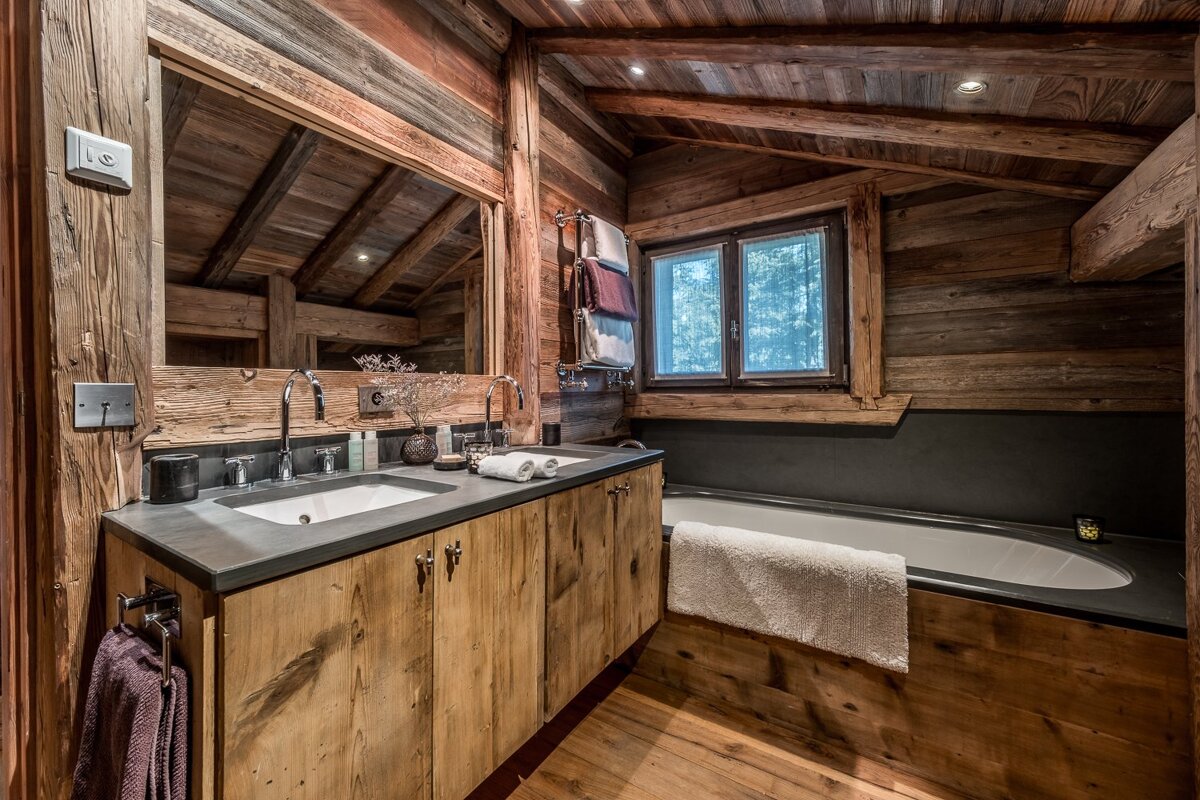Rustic wooden bathroom featuring a double vanity, bathtub by a window, and exposed wood walls and ceiling.