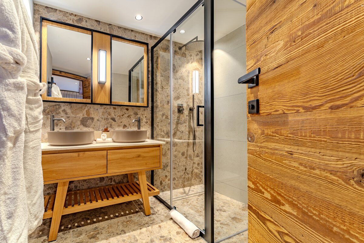 Contemporary bathroom with a double wood vanity, grey vessel sinks, and a glass shower. Features warm stone and wood textures, with a white robe hanging.
