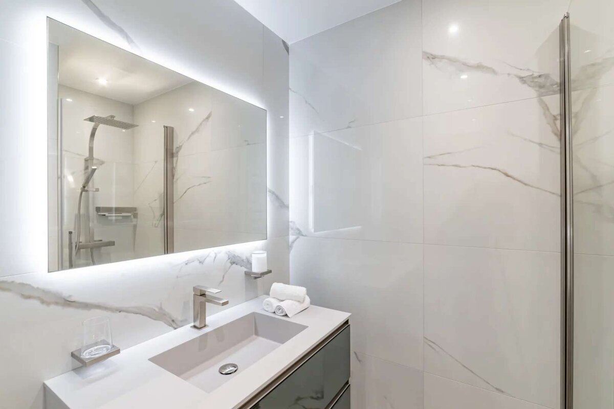 A modern, minimalist bathroom with white marble-look tiles, a vanity, and an illuminated mirror reflecting a shower. The space is clean and bright.