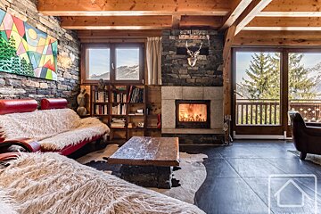 Cozy chalet living room with a lit stone fireplace, wooden beams, and stunning mountain views from large windows. Features comfy couches with fur throws.