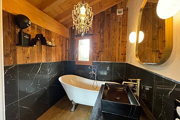 Luxurious bathroom with a clawfoot tub, black marble tiles, wooden walls/ceiling, and a crystal chandelier, featuring gold accents.