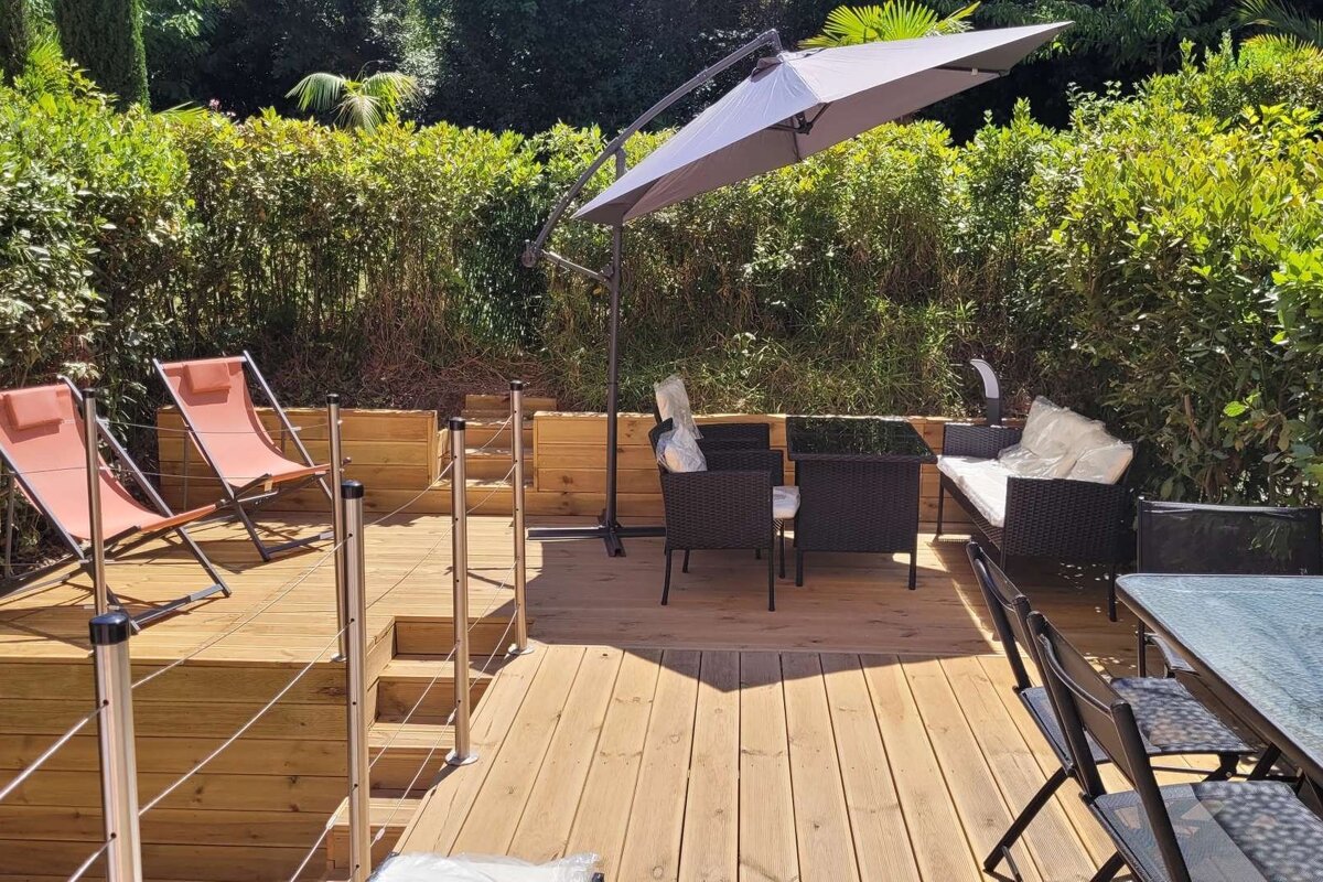 A sunny wooden deck with various outdoor furniture, including lounge chairs, a dining set, and sofa, surrounded by lush green foliage.