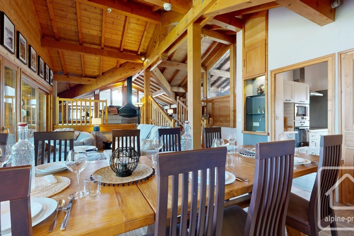 A warm, open-plan wooden chalet interior with a dining table, living area with fireplace, and kitchen visible in the background.