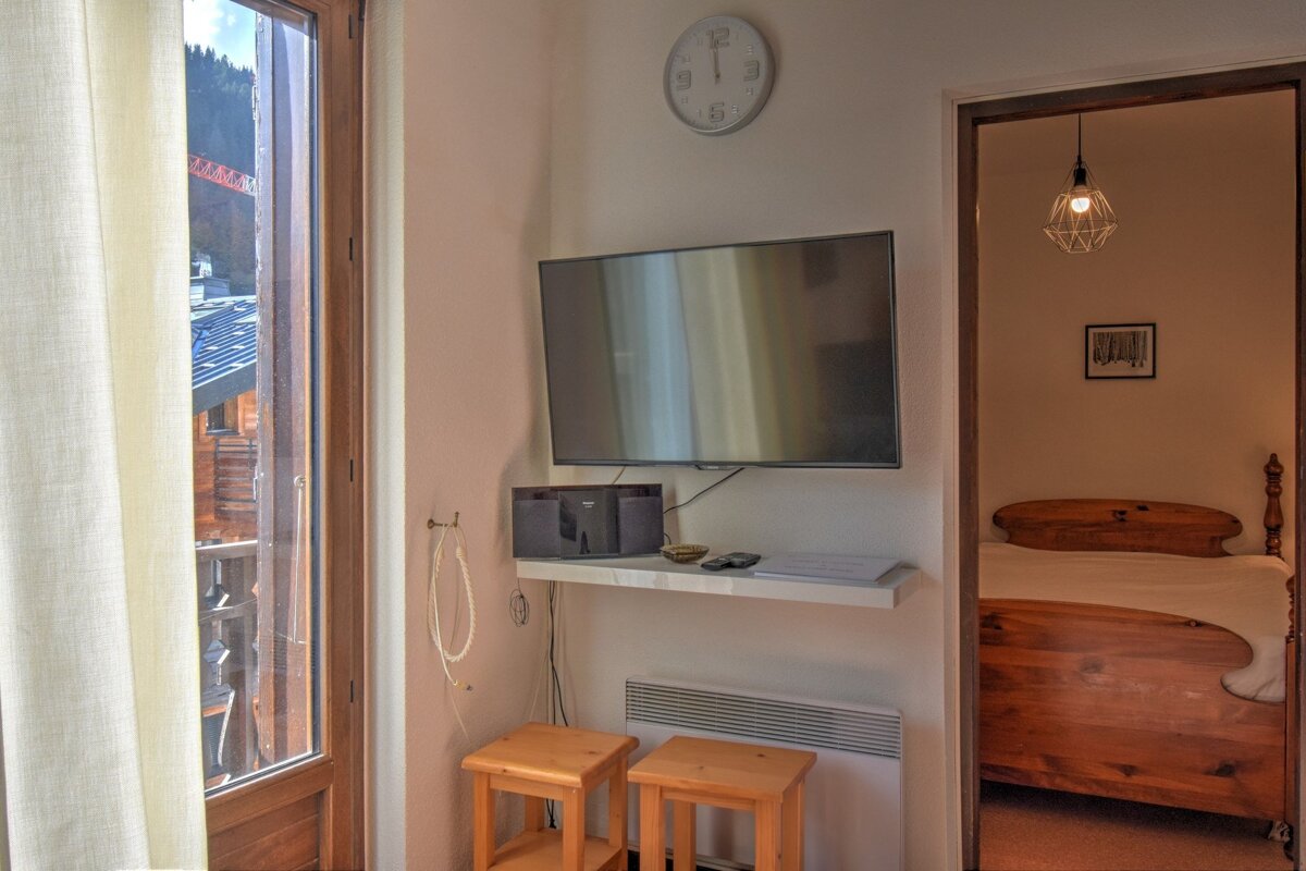 A room with a wall-mounted TV, small stools, and a window overlooking mountains. An open doorway reveals a bedroom with a wooden bed.