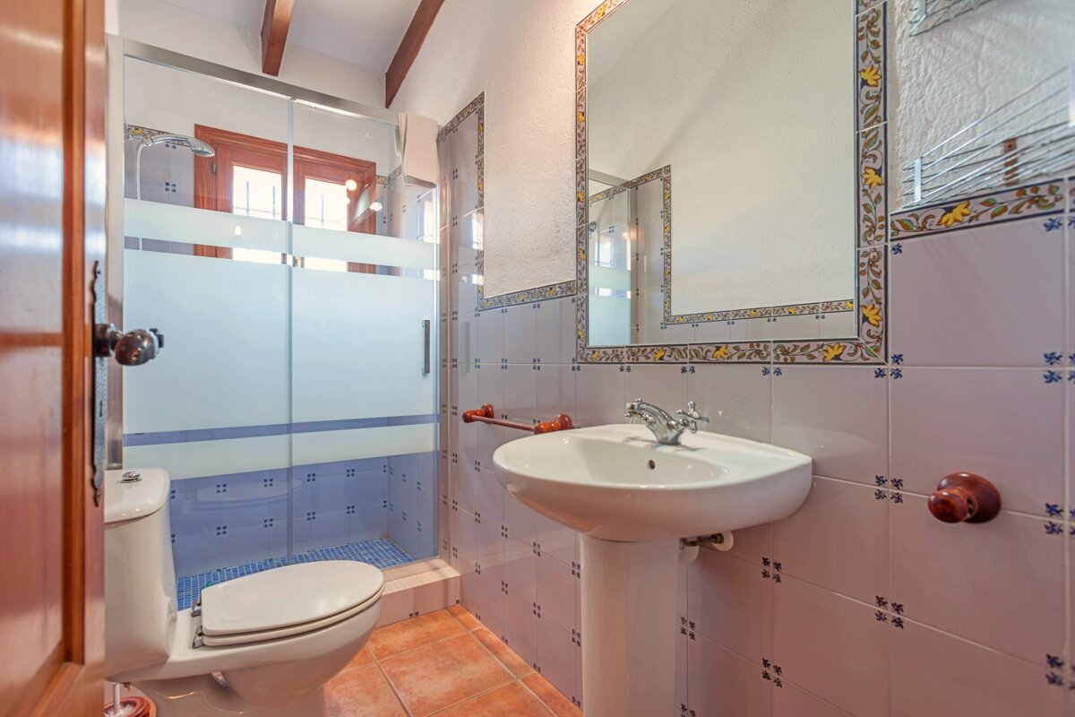 A bright bathroom with a frosted glass shower, toilet, pedestal sink, and mirror. It features decorative blue and light pink tiled walls and terracotta floor.