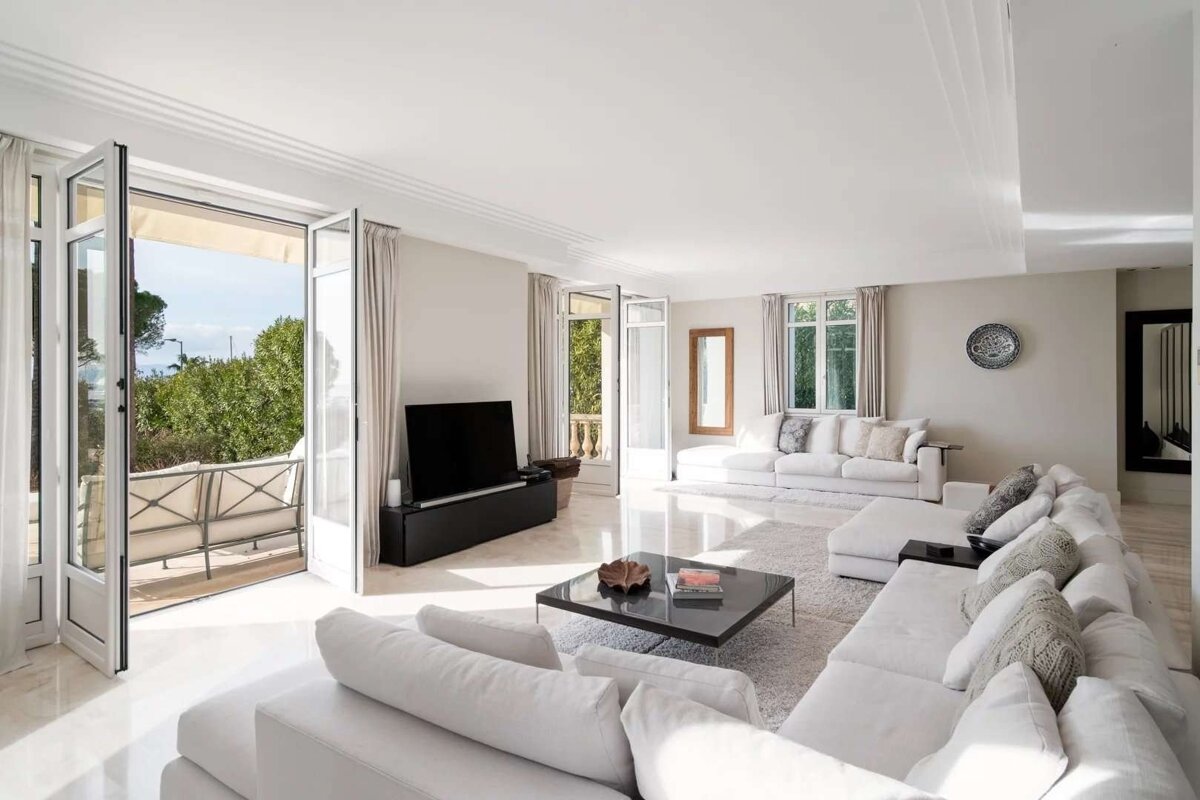 A bright, modern living room with multiple white sectional sofas, a black coffee table, and an open balcony with lush greenery visible through large glass doors.