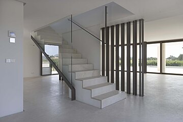 Modern, minimalist room with light concrete floors, a white staircase with glass railing, and dark slatted partition. Large windows offer vineyard views.