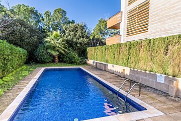 A rectangular blue swimming pool with steps, surrounded by lush green hedges and trees, next to a modern building under a clear blue sky.