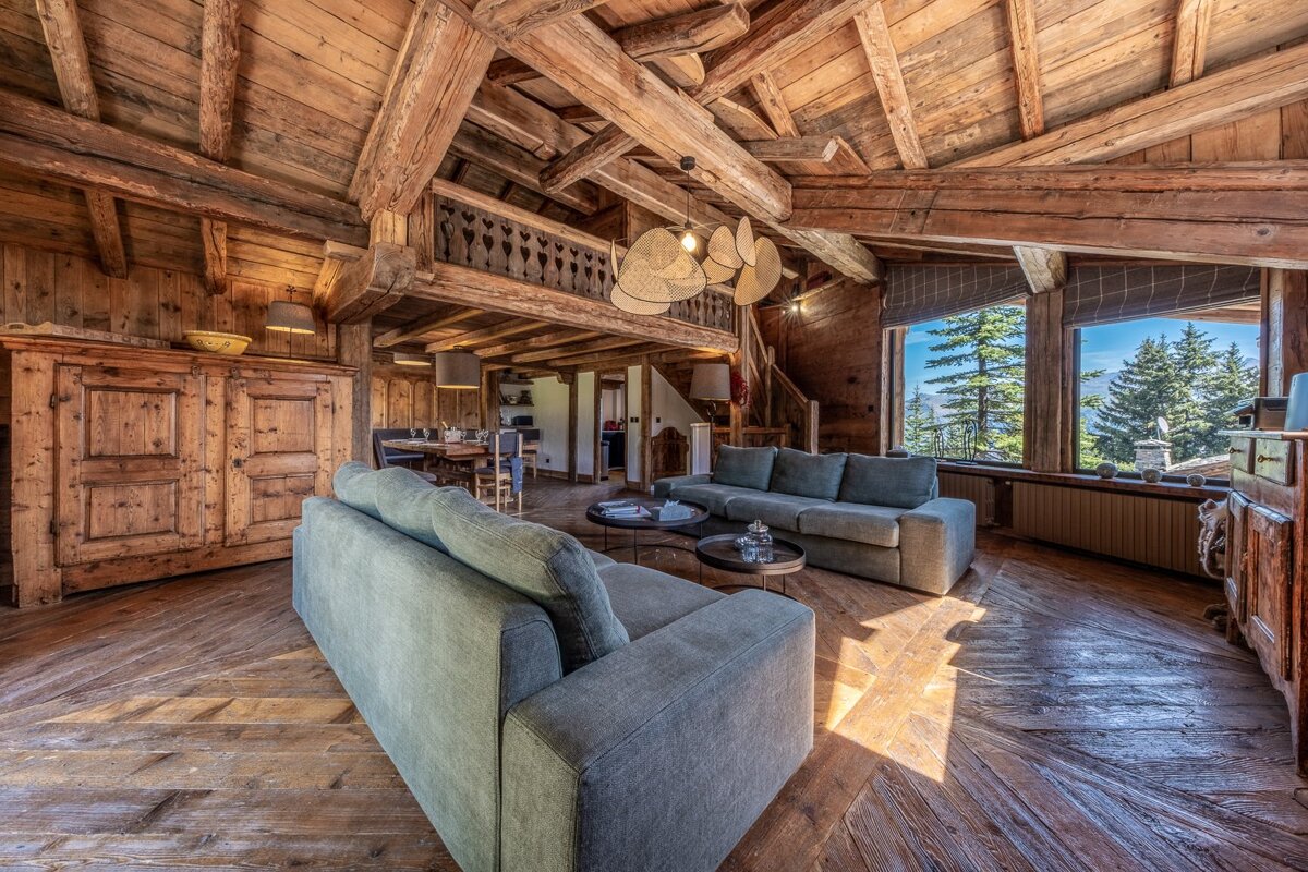 A spacious, rustic wooden chalet living room with modern gray sofas, high beamed ceilings, and large windows showcasing a scenic mountain forest view.