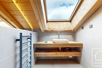 A stylish attic bathroom with a wooden vanity, vessel sink, gray tiled walls, towel warmer, and a large skylight showing the sky.