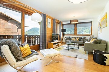 Modern chalet living room featuring wood-paneled walls, comfortable sofas, a chic white armchair, and large windows with mountain views.