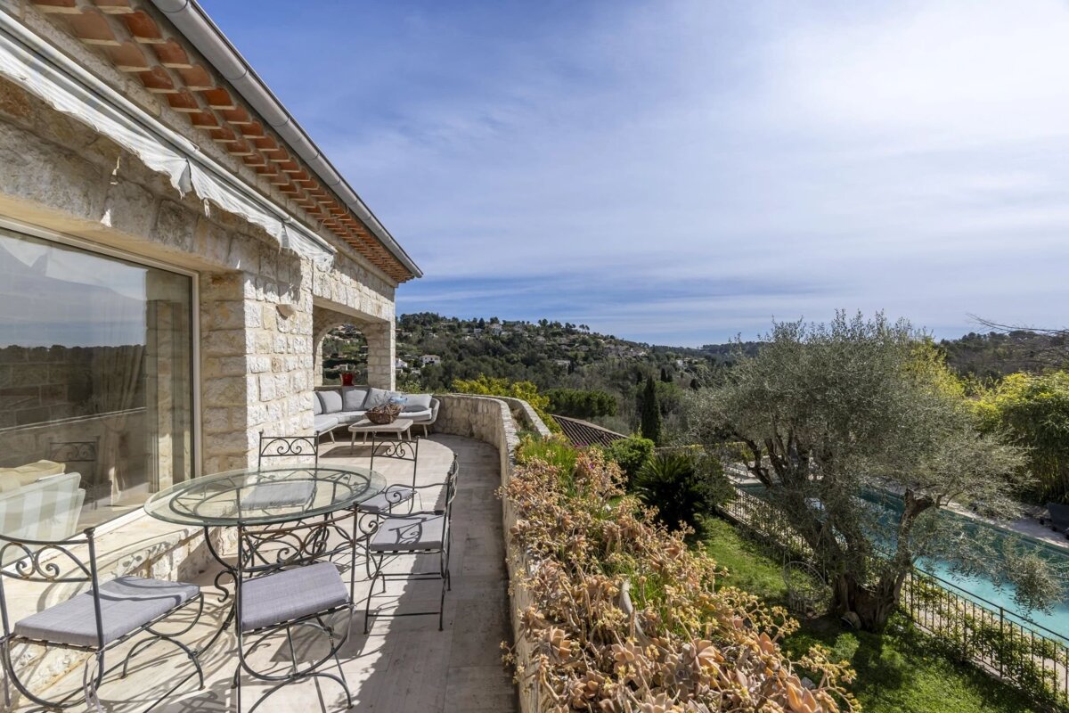 Sunny stone villa terrace with outdoor seating, a sparkling pool, and panoramic views of lush green hills and distant sky.