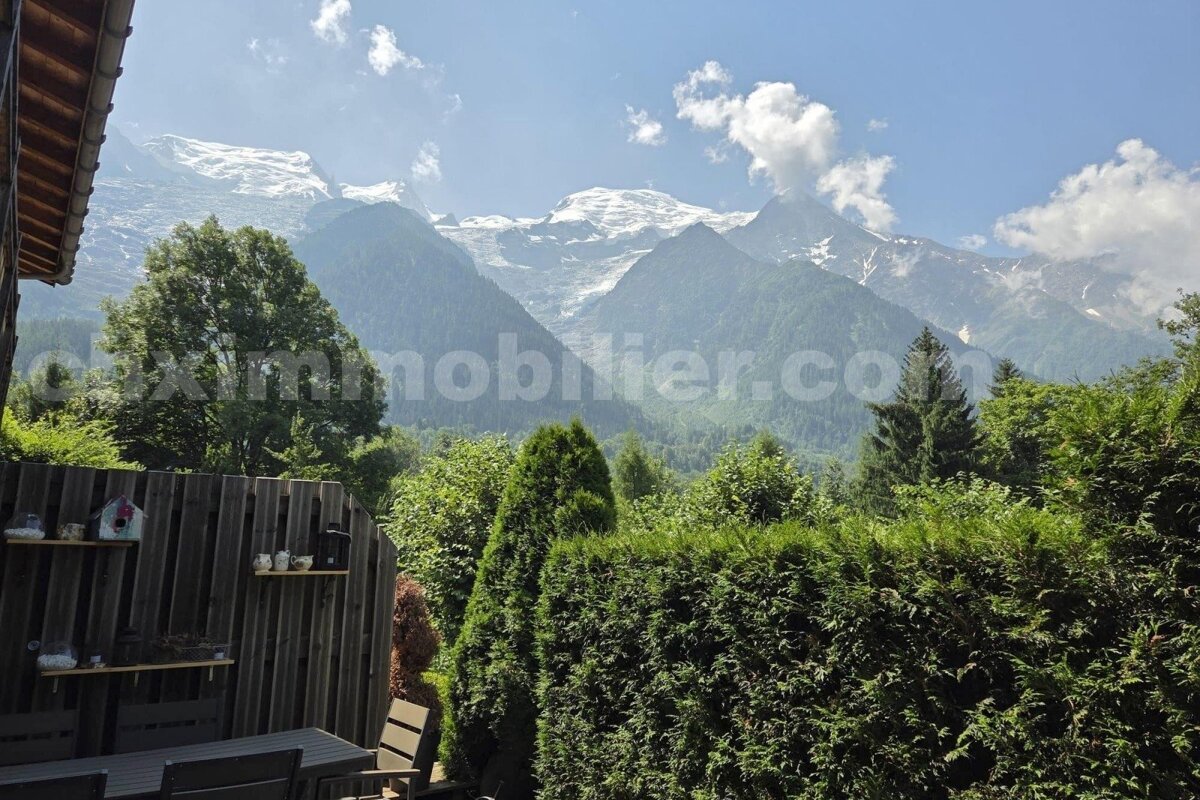 Majestic snow-capped mountains rise above a green valley, viewed from a private garden/patio with hedges and trees. Bright blue sky.