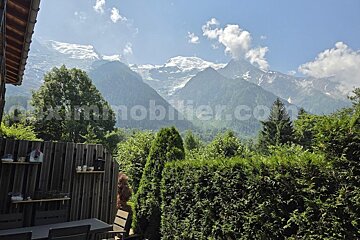 Majestic snow-capped mountains rise above a green valley, viewed from a private garden/patio with hedges and trees. Bright blue sky.