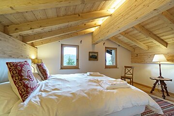 Cozy attic bedroom with slanted wooden ceilings and beams, white bedding with red patterned pillows, and windows with mountain views.