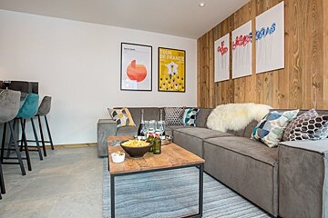 Cozy modern living room with a grey sectional, wooden coffee table, wood-paneled accent wall, and colorful wall art.
