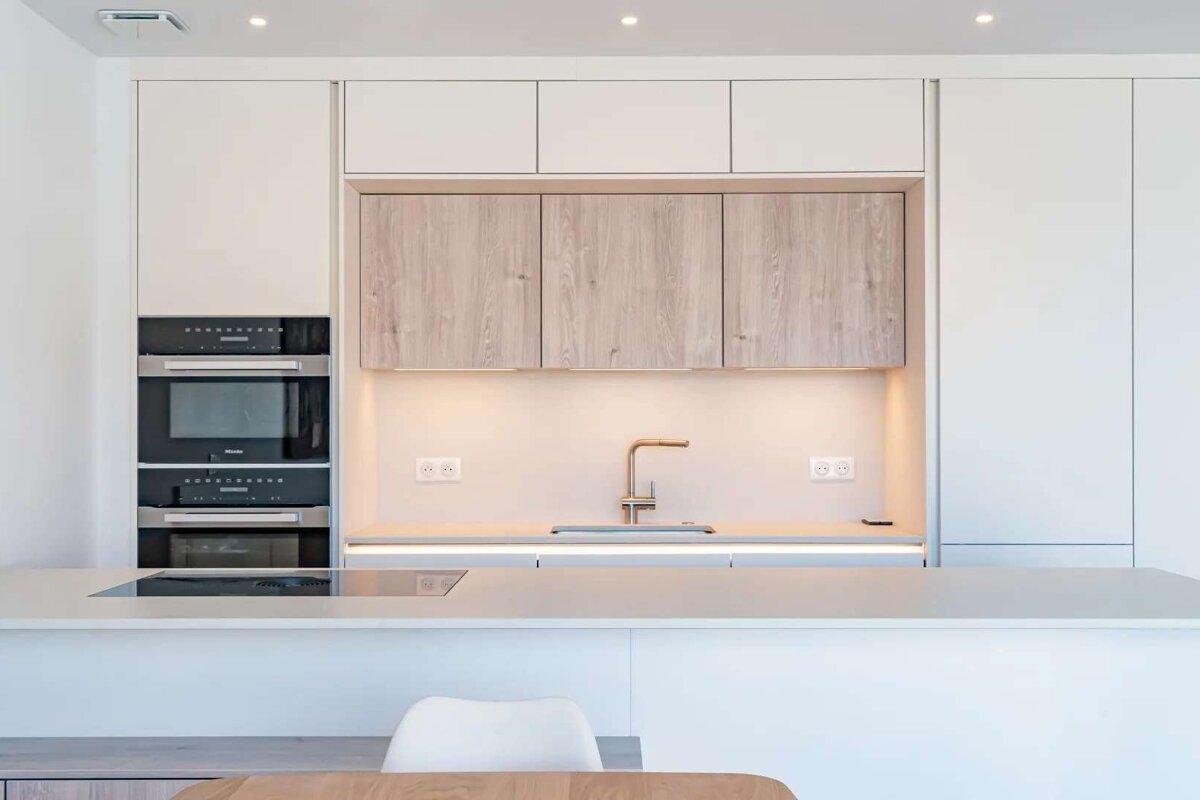 A bright, modern kitchen with white and light wood cabinets, built-in ovens, a large island with a sink, and warm under-cabinet lighting.
