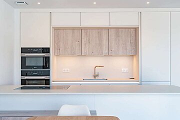 A sleek, minimalist kitchen featuring white and light wood cabinetry, integrated appliances, an island with a stovetop, and a stylish sink.