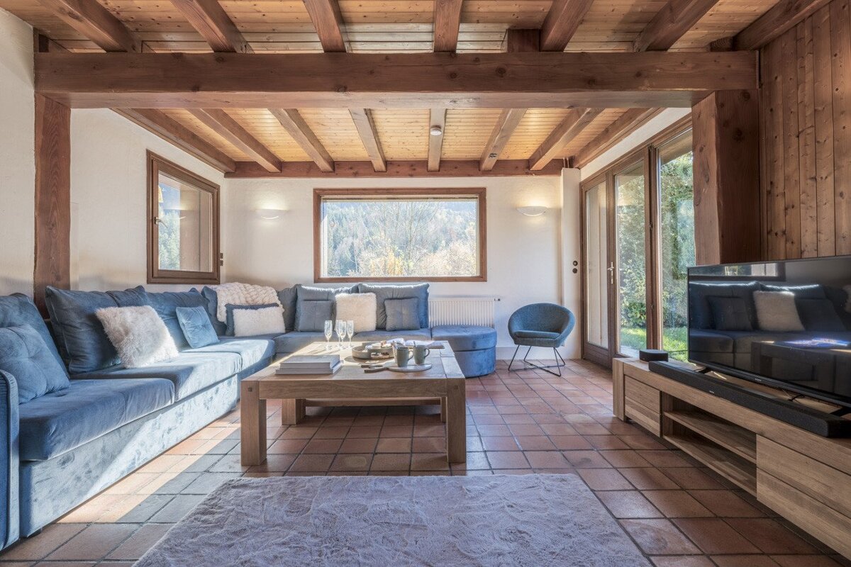 Cozy living room with a blue sectional sofa, exposed wooden beams, tiled floor, and large windows showcasing a nature view. Modern rustic comfort.
