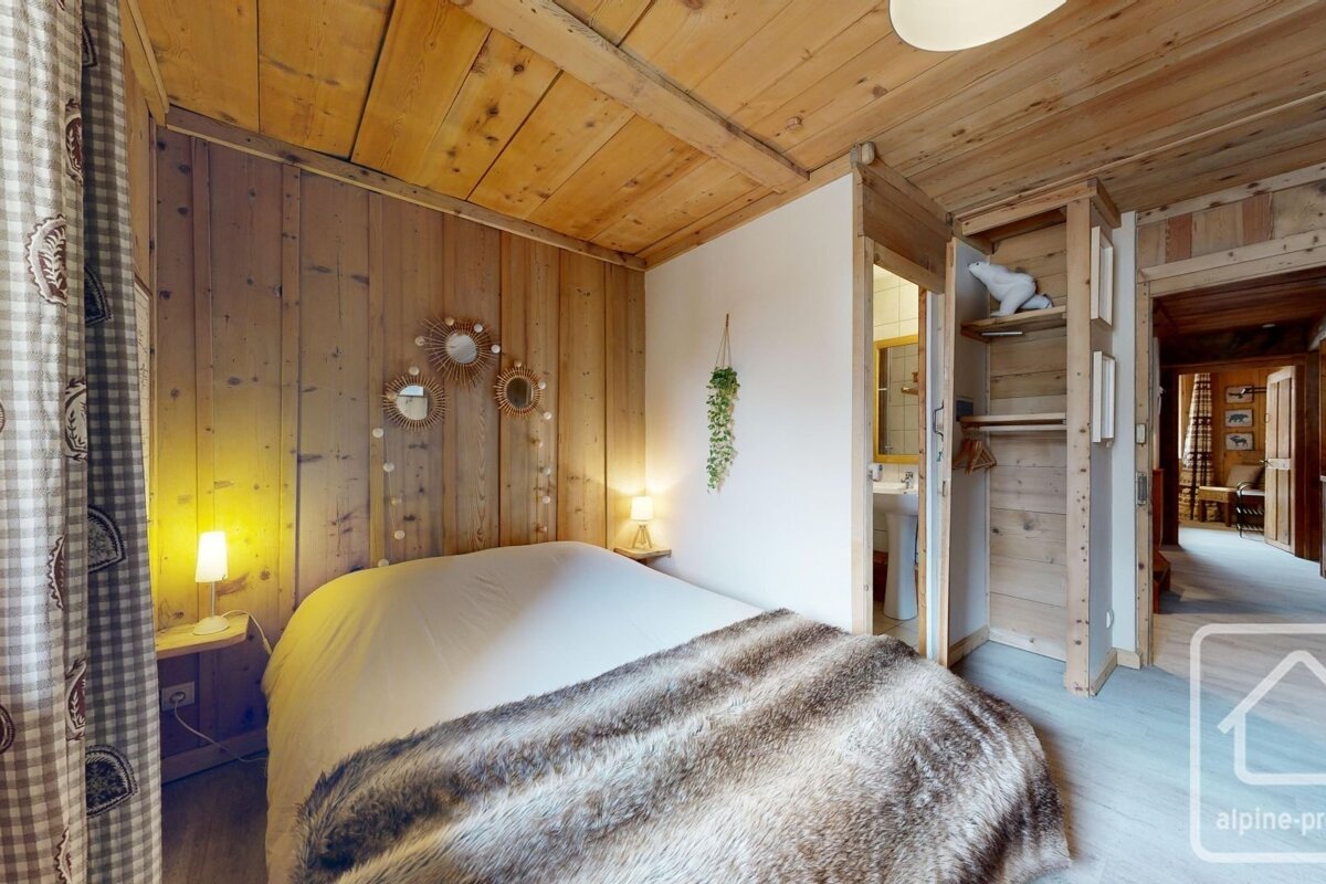A cozy, rustic bedroom with wood-paneled walls/ceiling, a bed with a furry throw, and a bedside lamp. A bathroom and hallway are visible.