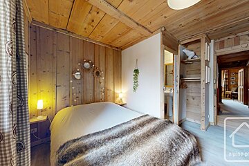Cozy, wood-paneled chalet bedroom with a faux fur throw, decorative mirrors, open wardrobe, and a peek into the bathroom/hallway.