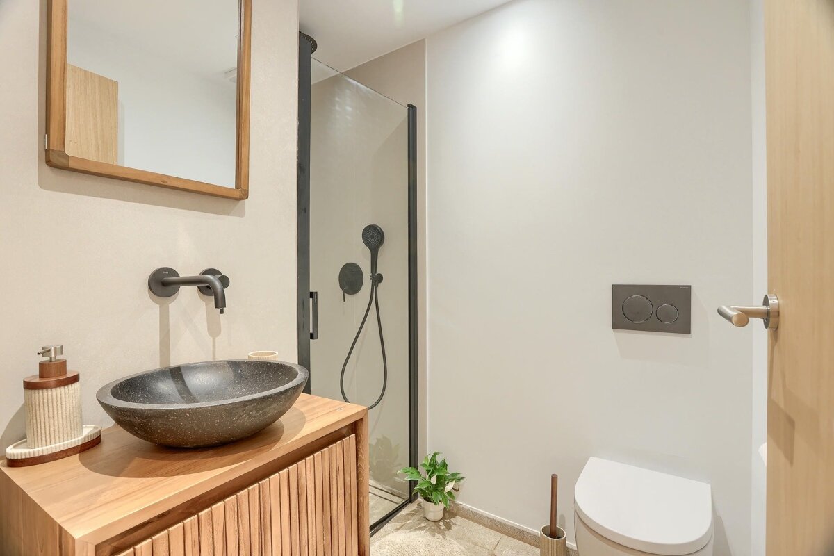 A modern bathroom features a wooden vanity with a stone sink, black fixtures, a glass shower, and a white toilet. Neutral tones create a clean, contemporary look.