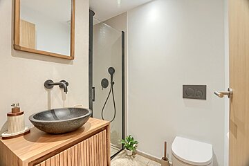 A modern bathroom features a wooden vanity with a stone sink, black fixtures, a glass shower, and a white toilet. Neutral tones create a clean, contemporary look.