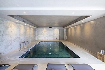 A luxurious indoor swimming pool with dark water, textured grey walls, subtle lighting, and poolside lounge chairs.