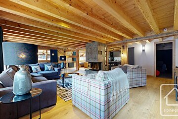 Spacious chalet living room featuring a warm wood-beamed ceiling, stone fireplace, blue sectional sofa, and plaid armchairs.
