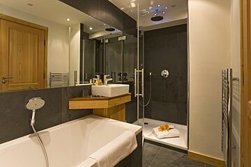 A sleek, modern bathroom featuring a white bathtub, glass shower with rain head, and a vessel sink on a wooden counter, all against dark grey tiles.