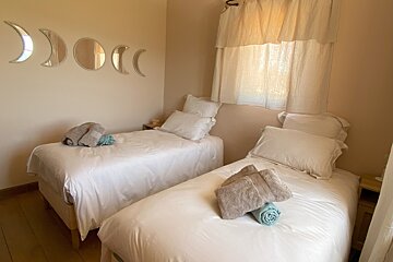 A light-colored twin bedroom features two beds with white linen and towels. Moon phase mirrors adorn the wall, and a window has sheer curtains.