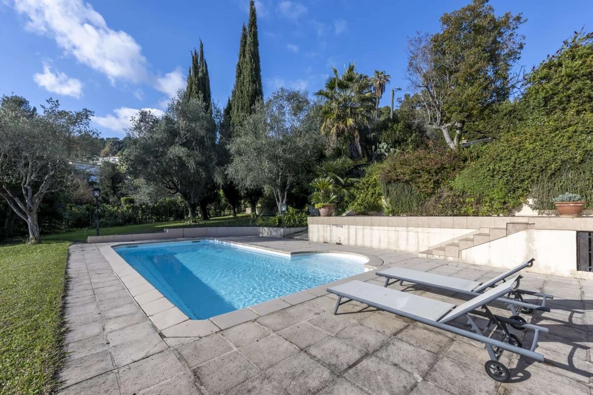 A sunny outdoor swimming pool with a stone patio and two lounge chairs, surrounded by lush green trees and a hillside under a blue sky.