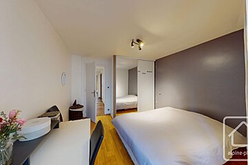 A bedroom featuring a large white bed, gray wall, wooden floor, sliding mirror closet, and a desk on the left.