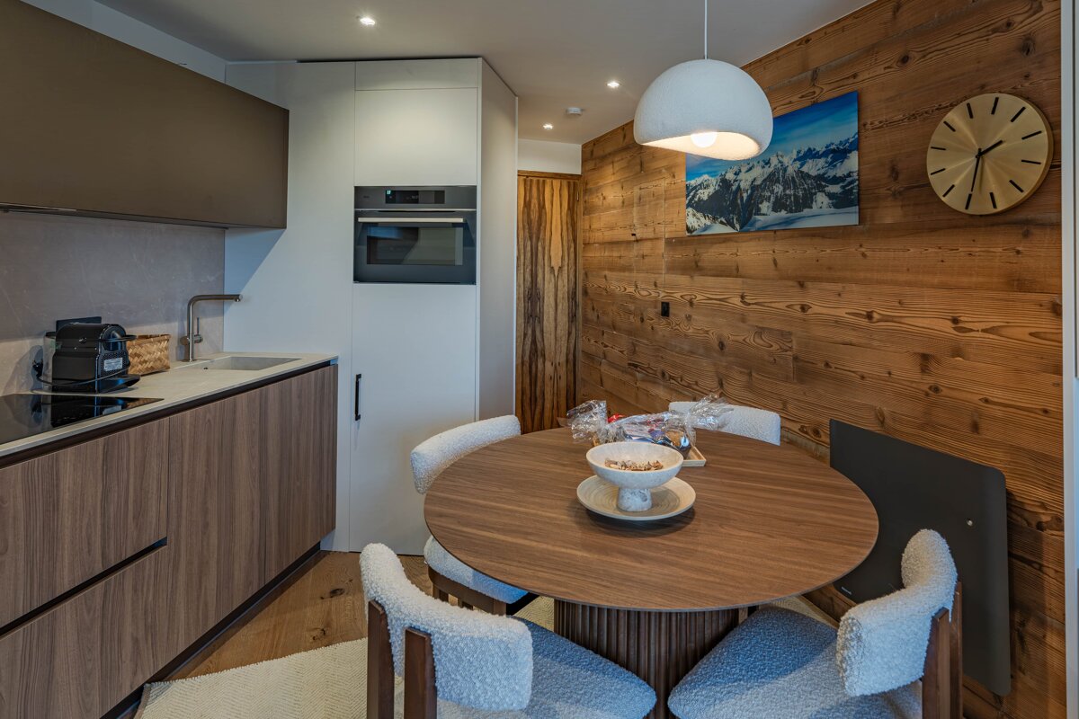 A modern kitchen and dining area featuring wood-grain cabinets, a round table with fluffy chairs, and a rustic wooden wall with mountain art.