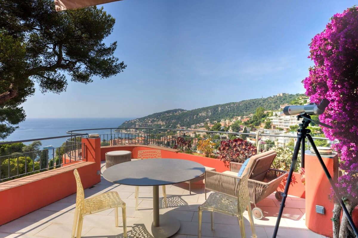A sunny terrace with outdoor seating, a telescope, and vibrant pink flowers, offering a panoramic view of the blue sea, coastal towns, and green mountains.