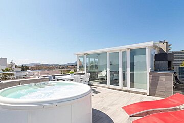 Sunny rooftop terrace with a bubbling hot tub, glass-enclosed room, outdoor dining area, red lounge chairs, and distant mountains.
