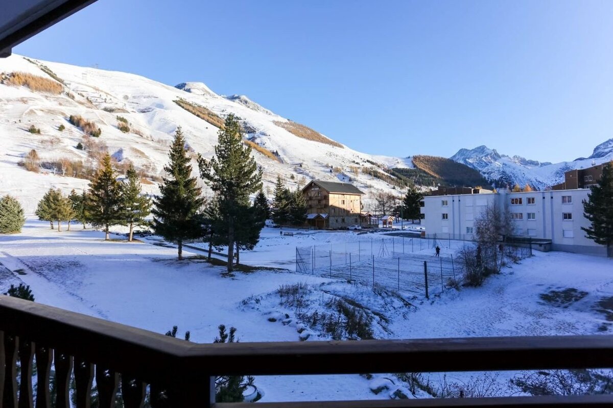 Balcony view of a snow-covered mountain resort with pine trees, buildings, and majestic peaks under a clear blue sky.