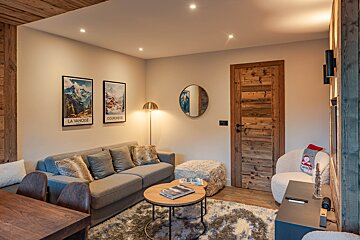 Cozy chalet living room featuring a grey sofa with plush pillows, wooden tables, a fluffy rug, and framed alpine travel posters.