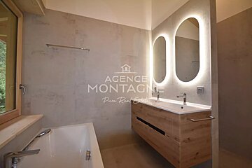 A modern bathroom featuring a wooden vanity with two illuminated oval mirrors, concrete-like walls, and a white bathtub.