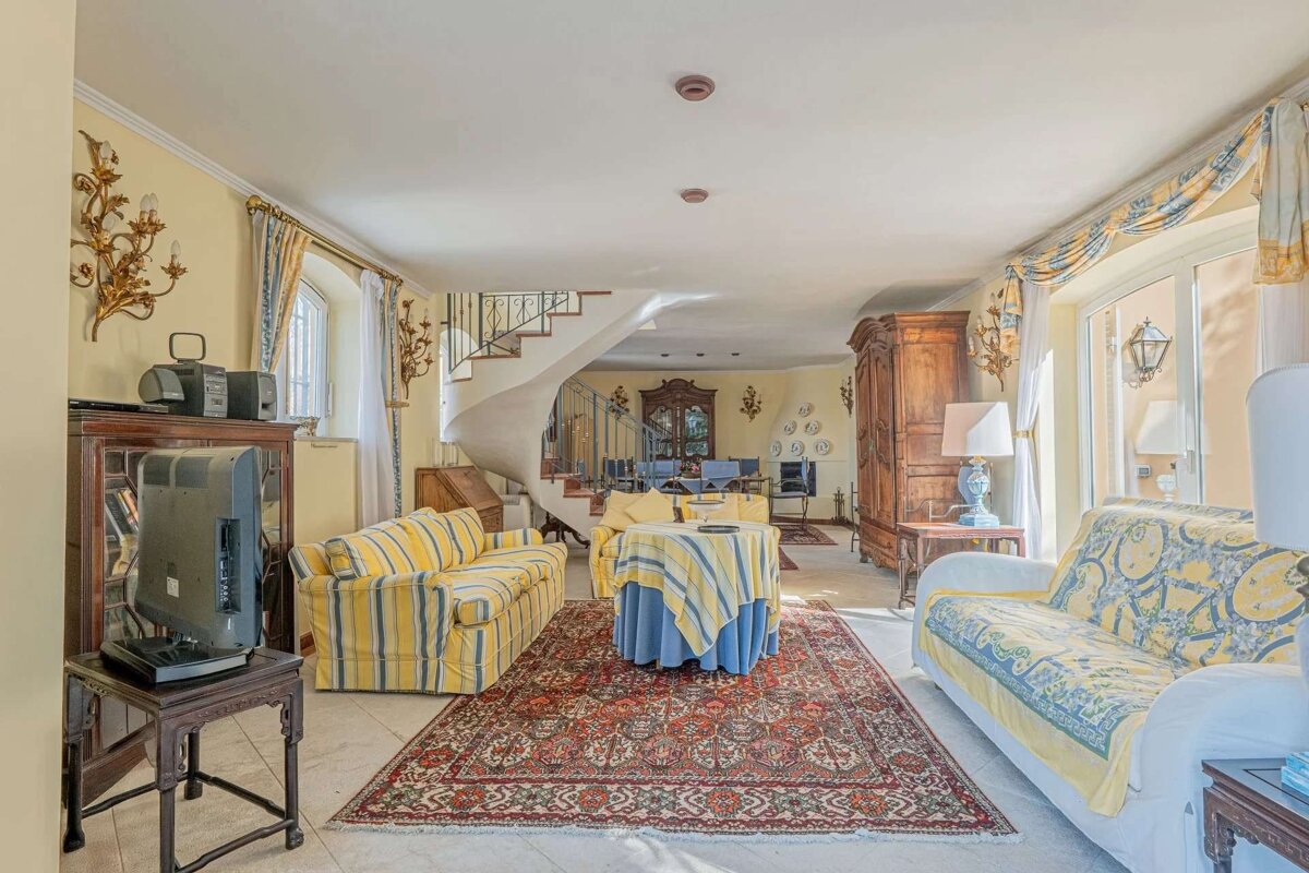 Bright, traditional living room with yellow walls, patterned sofas, and a red rug. Features a spiral staircase, ornate decor, vintage TV/stereo, and wooden armoire.